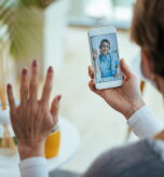 Close-up of woman greeting her doctor while using smart phone and having video call.  Focus is on female doctor on touchscreen.